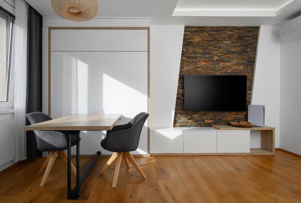 Jídelní Stůl Se Do úzké Kuchyně Nevešel. Podlaha Z Masivního Dřeva Příjemně Kontrastuje S Lesklou Plochou Stěny Za Stolem. Barevně S Ní Ladí Obklad Za Televizorem.